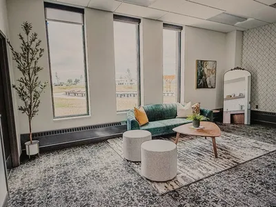 Modern lounge area with teal sofa, oval coffee table, two ottomans, and tall windows overlooking a flat landscape