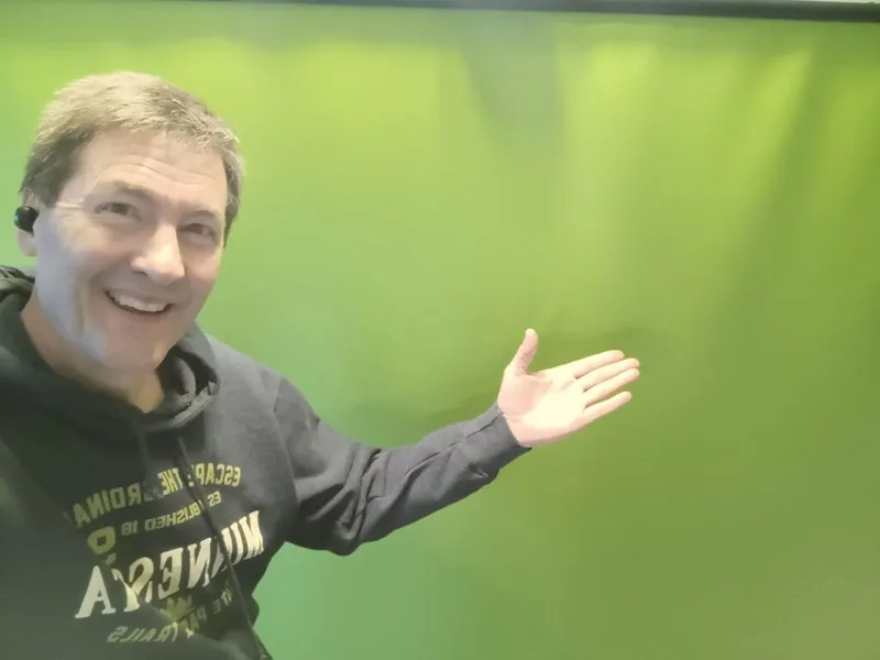 Smiling man in a dark hoodie gesturing toward a green screen backdrop