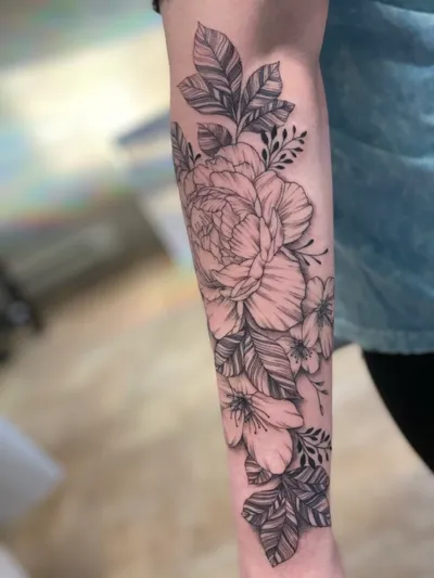 Forearm tattoo featuring a large peony and smaller flowers with detailed leaves in black and grey linework