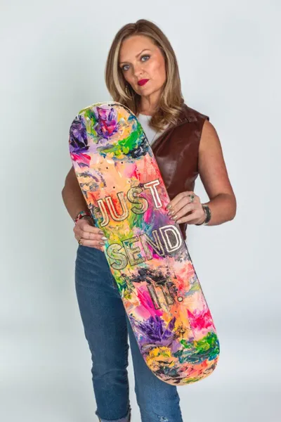 Woman in brown leather vest holding a colorfully painted skateboard reading 'Just Send It
