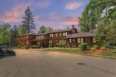 Large rustic wood-sided lodge surrounded by tall pines under a pink evening sky, with golf carts parked out front