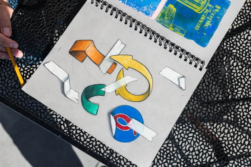 Hand holding a pencil beside an open sketchbook with colorful arrow and logo sketches