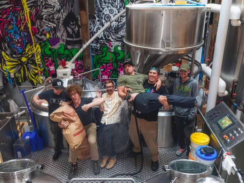 Group of brewery workers posing playfully among stainless steel tanks with bold colorful murals behind them