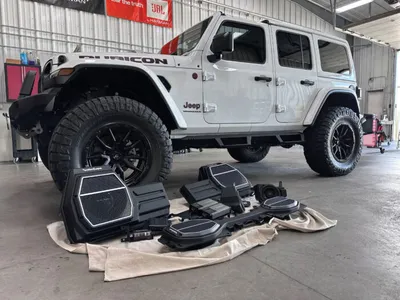 White Jeep Rubicon lifted on large black rims with audio equipment laid out on a drop cloth in a garage