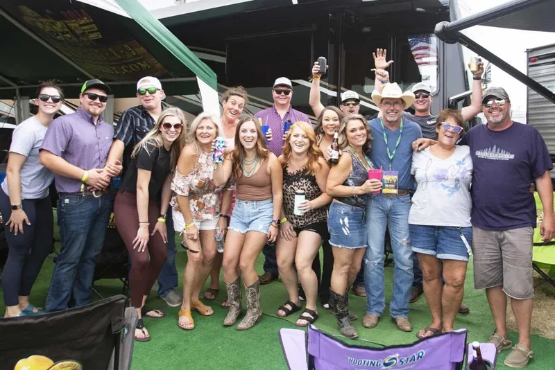 Large group of festivalgoers posing and smiling outdoors near an RV at an outdoor music festival