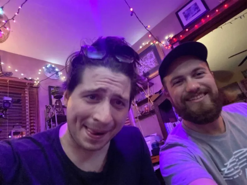 Two men smiling for a selfie in a dimly lit room with string lights