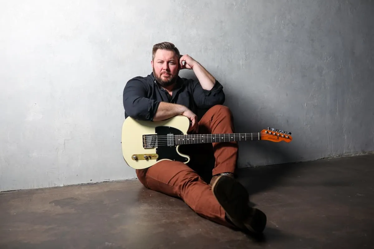 Bearded man in dark shirt and rust pants sitting on floor holding a cream electric guitar