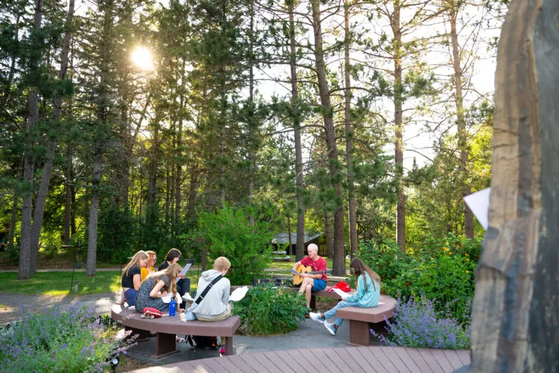 Small group of young people seated on outdoor benches listening to a man play guitar among tall pine trees