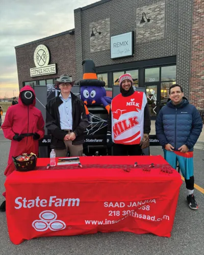 Four people in costumes stand behind a red State Farm branded table outside a coffee house