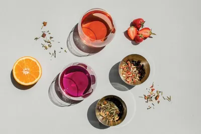 Two colorful drinks, dried herb blends in small bowls, a halved orange, and fresh strawberries on a light surface