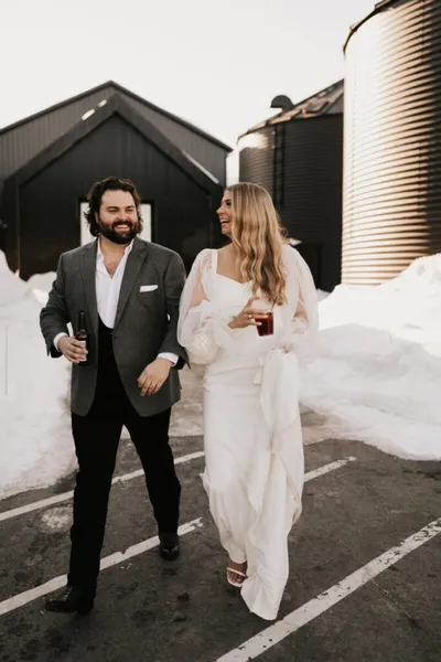 Smiling couple in wedding attire walking outdoors near a dark barn and grain silo in winter snow