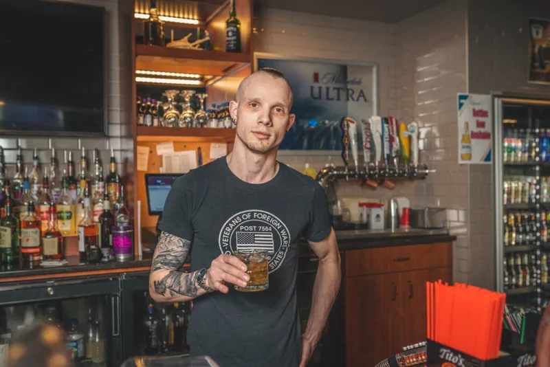 Tattooed bartender holding a whiskey drink behind a well-stocked bar