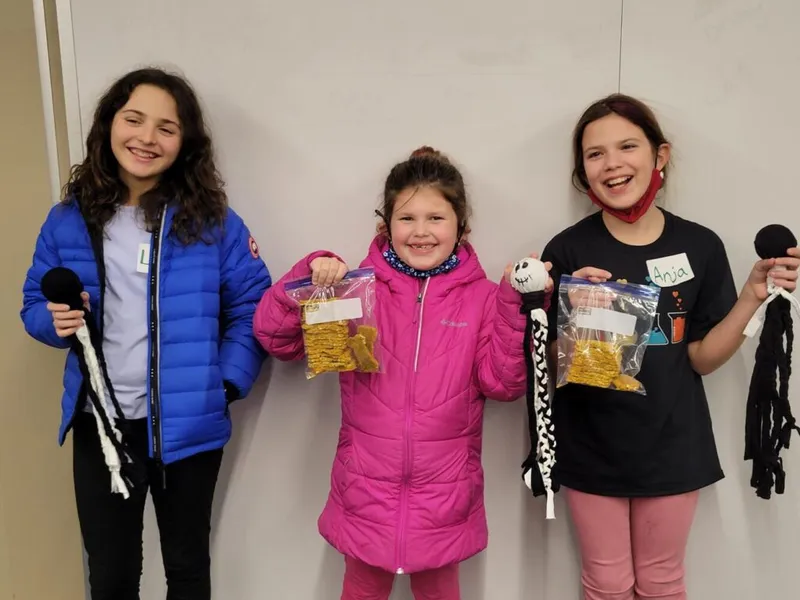 Three smiling girls holding handmade yarn crafts and bags of snacks