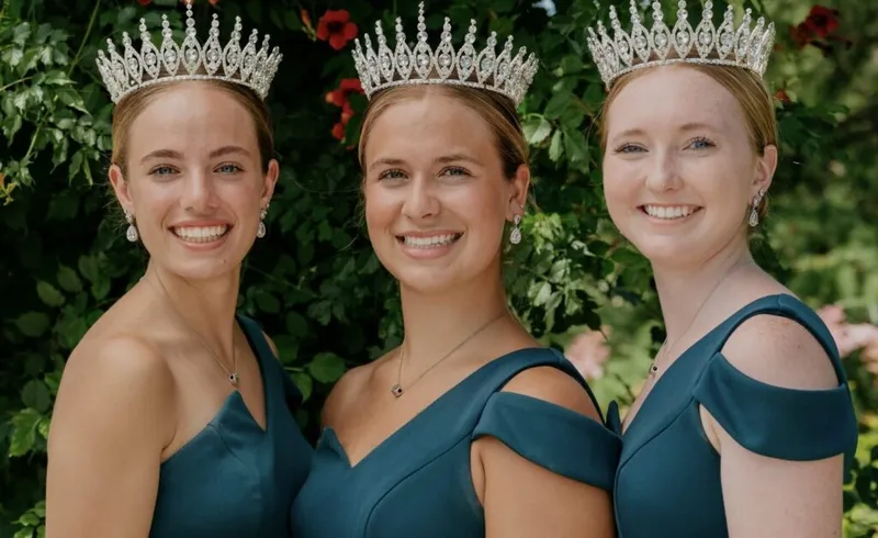 Three smiling women wearing tiaras and matching teal off-shoulder gowns posed before green foliage