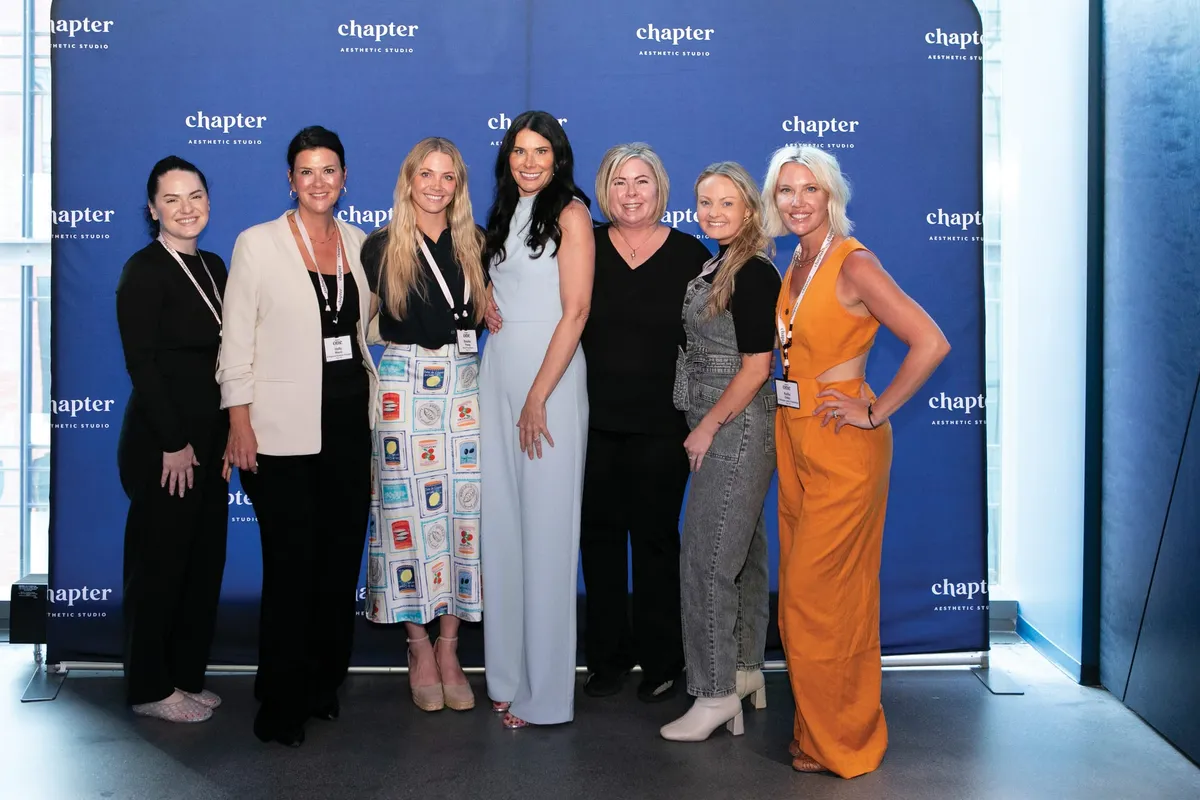 Seven women posing together in front of a Chapter Aesthetic Studio branded backdrop