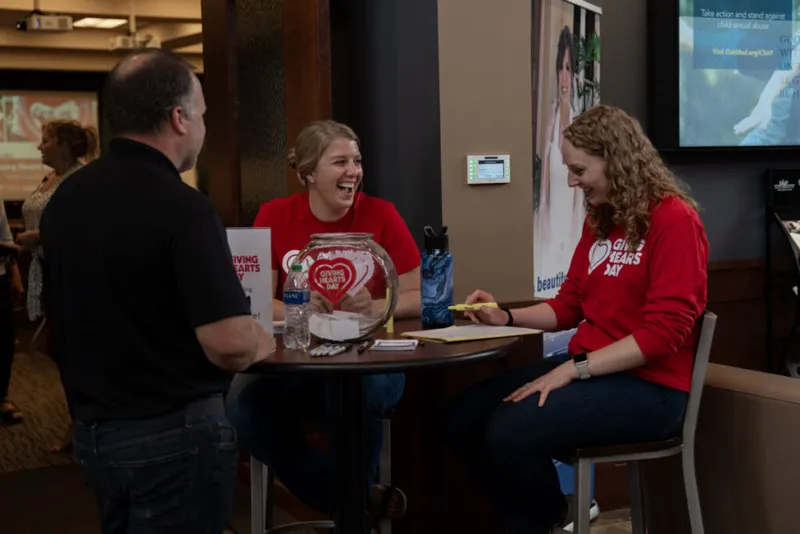 Two smiling volunteers in red Giving Hearts Day shirts chat with a visitor at a round table