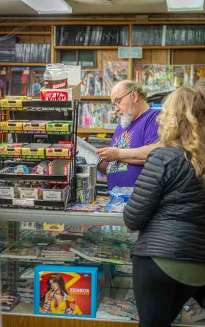 Bearded man in purple tee reviews comics behind a cluttered shop counter with a customer