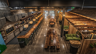 Aerial view of a spacious brewery interior with long wooden communal tables, bar seating, and warm industrial décor