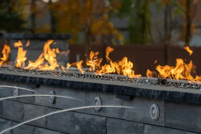 Outdoor gas fire pit with orange flames and glass beads, autumn trees in background