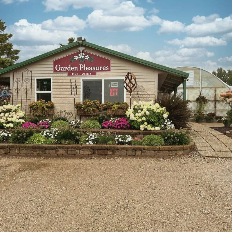 Garden Pleasures Est. 2001 garden center storefront with colorful flower beds and a greenhouse