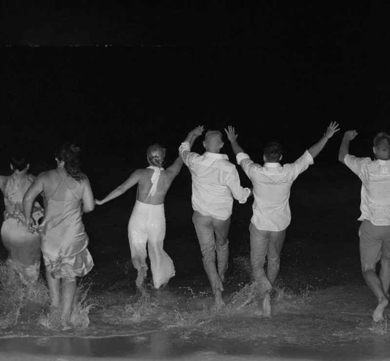 Group in formal attire running into ocean waves at night, arms raised