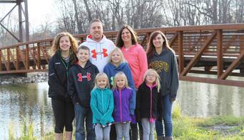 A smiling family of nine poses outdoors near a wooden bridge over a pond