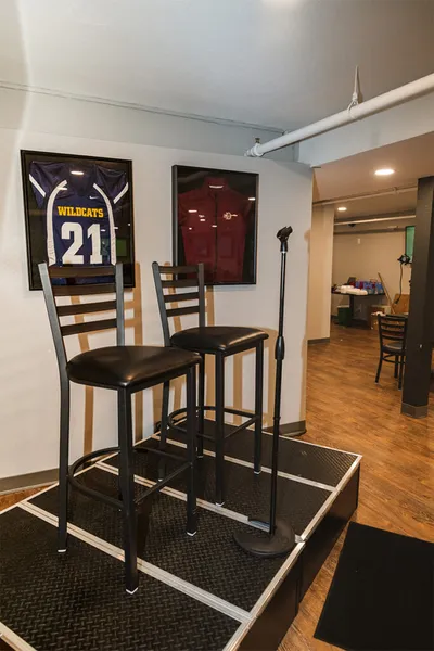 Two bar stools and a microphone stand on a small stage with framed sports jerseys on the wall behind