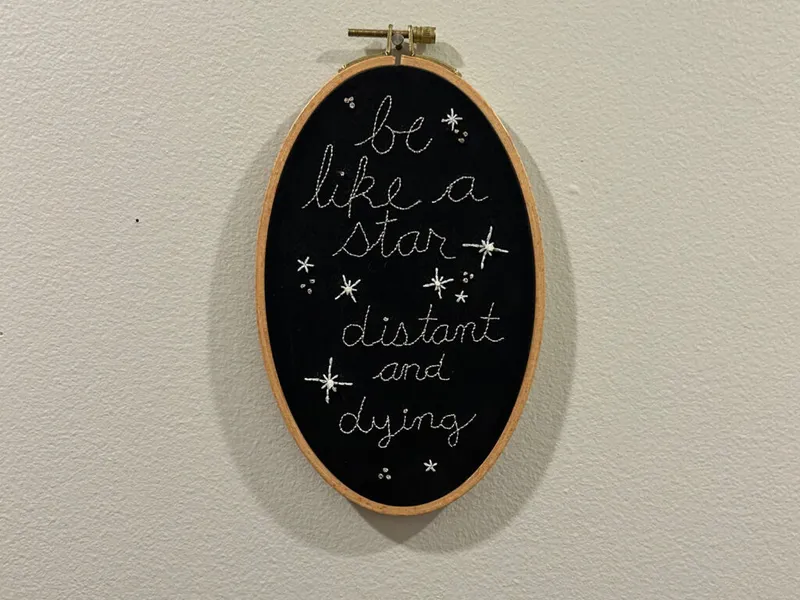 Oval embroidery hoop on white wall with white stitched text "be like a star, distant and dying" on black fabric