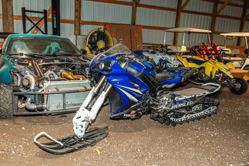 Blue Yamaha snowmobile bike conversion parked beside a modified car with exposed engine and a yellow ATV inside a barn