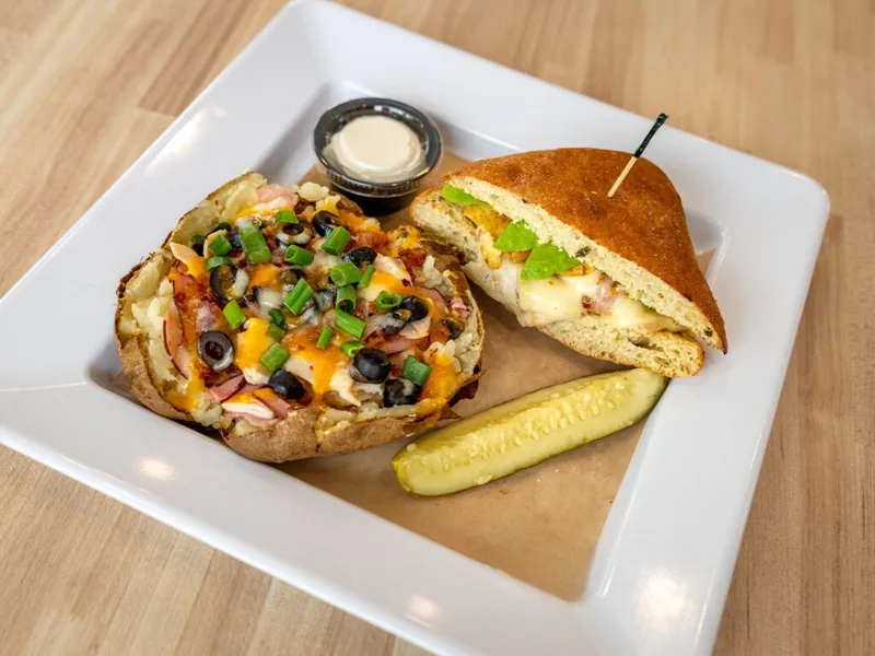 Open-faced loaded bagel with olives, cheese, and green onions beside a sandwich half and pickle on a white plate