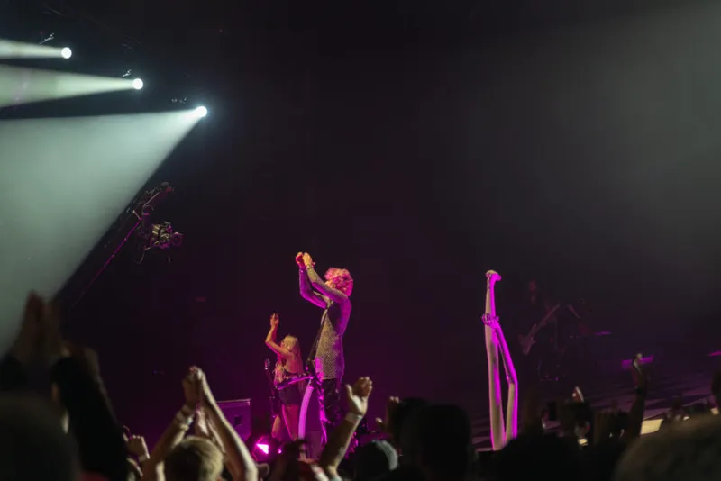 Performer in a glittery outfit raises arms on a purple-lit stage before a crowd
