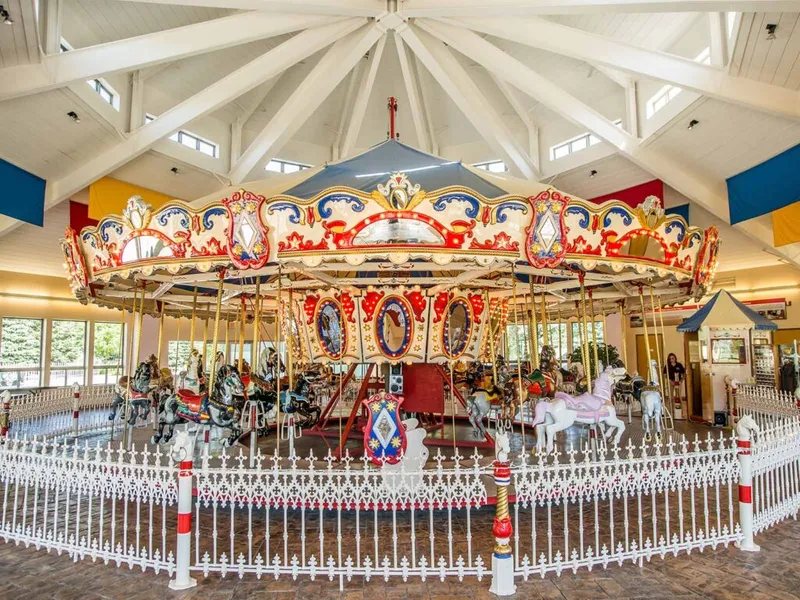 Ornate indoor carousel with red and gold canopy and white carved horses behind a decorative fence