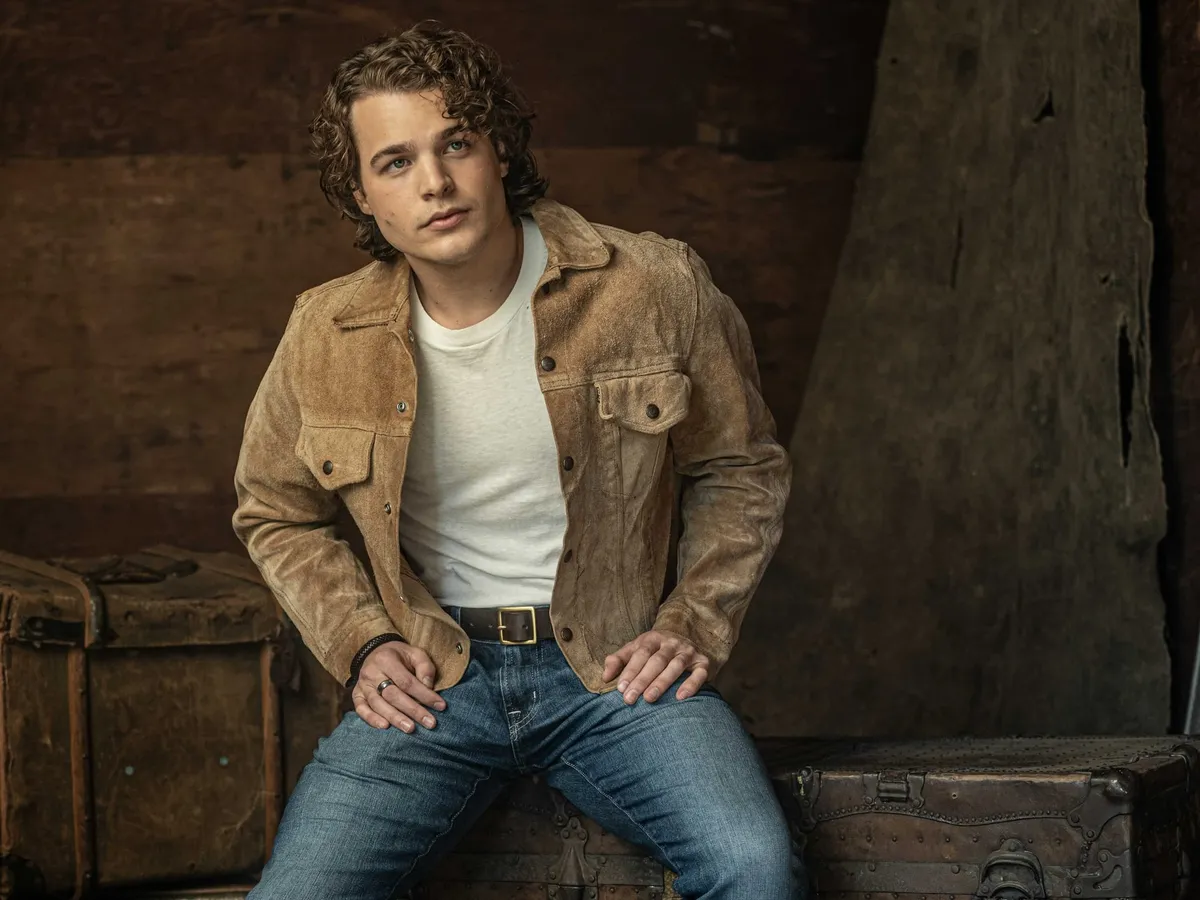 Young man with curly hair in a tan suede jacket and jeans seated on a vintage trunk