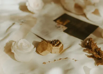 Gold hexagonal ring box with wedding bands on white silk fabric, near white roses and dried botanicals