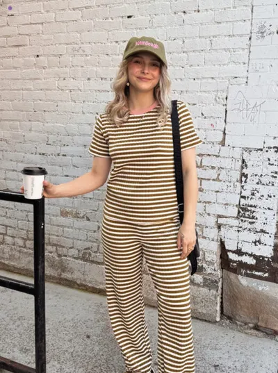 Woman in olive-striped matching set and green cap holds a coffee cup against a white brick wall