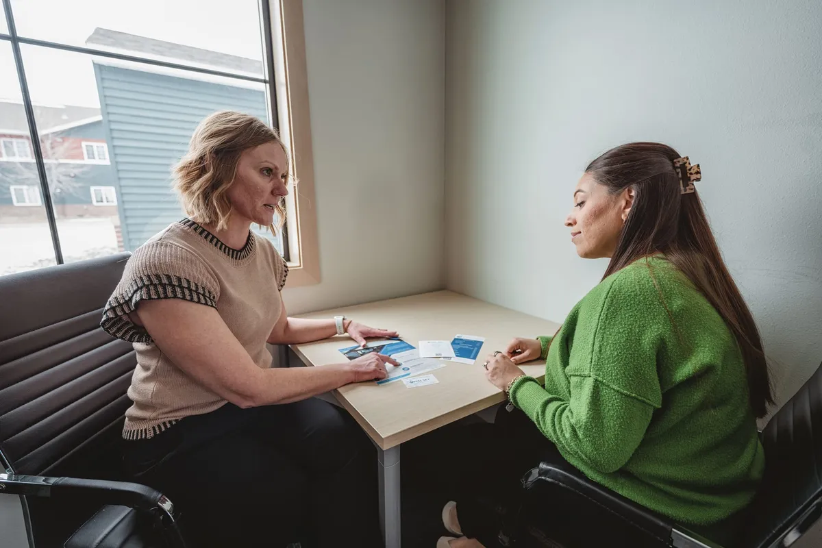 Two women reviewing blue pamphlets together at a small office table by a window