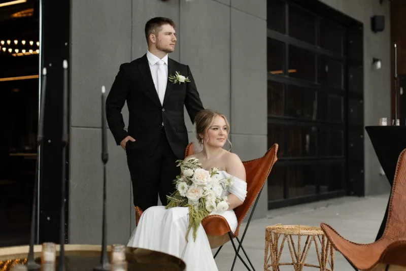 Bride seated in leather chair holding white bouquet, groom standing behind her in black suit
