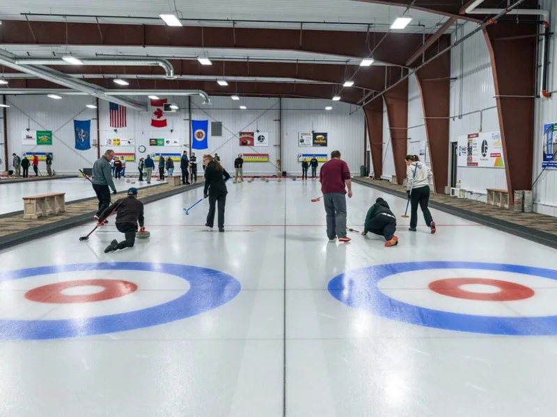Players sweep and deliver stones on two curling sheets inside an indoor rink with US and Canadian flags on the wall