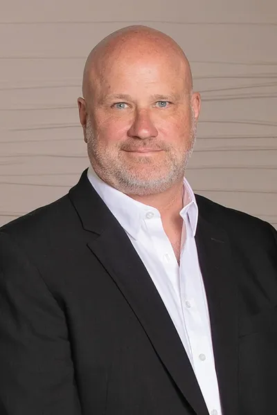 Bald middle-aged man with gray stubble wearing a black blazer and white shirt
