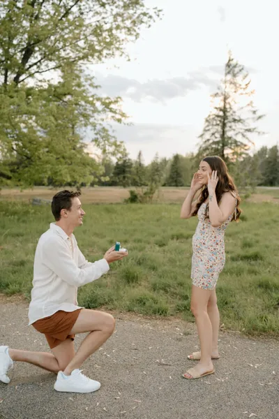Man kneeling on a park path holding a ring box toward a surprised woman in a floral dress