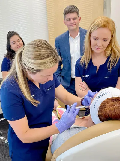 Two aestheticians in blue scrubs perform a facial treatment on a reclining client while two colleagues observe