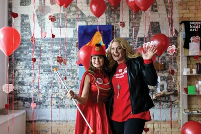 Two women in red outfits pose among red balloons and heart decorations
