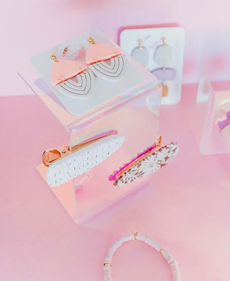 Pastel clay earrings and hair clips displayed on a clear acrylic cube against a pink background