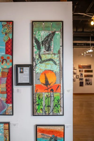 Tall framed mixed-media artwork with butterfly, orange sun, and botanical silhouettes hanging in a gallery