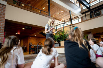 Woman addresses a group of young girls seated on the floor in a bright indoor atrium