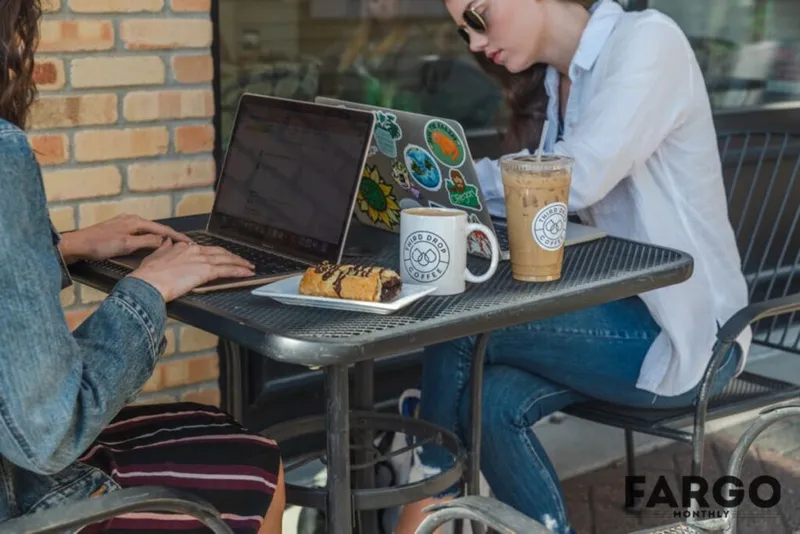 Two women at an outdoor metal café table with laptops, coffee, and a pastry
