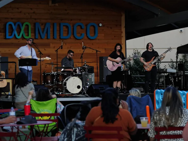 Four-piece band performing on an outdoor stage beneath a Midco sign before a seated audience