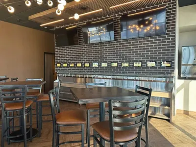 Restaurant dining area with high-top tables, metal chairs, brick accent wall, and mounted TVs