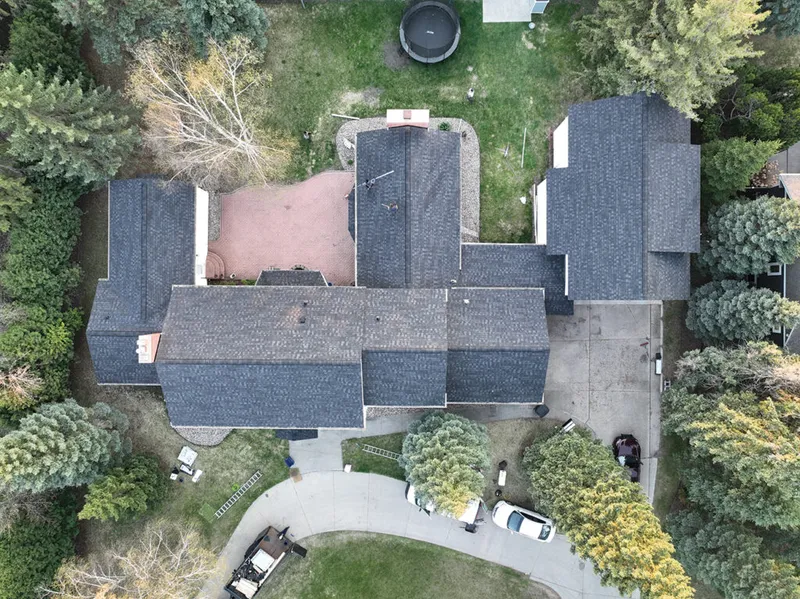 Aerial view of a large suburban home with dark gray shingled roof surrounded by trees