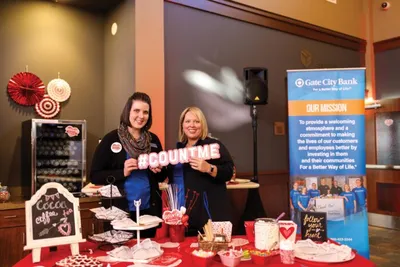 Two women smile behind a red-draped table holding a #COUNTME sign at a Gate City Bank event